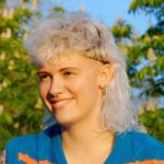 A young woman with a curly white blonde mullet haircut sitting outside on a sunny day.
