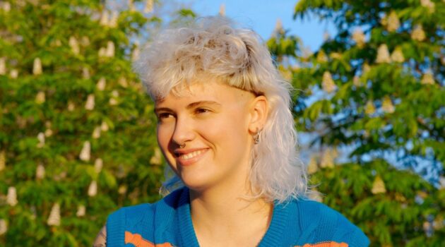 A young woman with a curly white blonde mullet haircut sitting outside on a sunny day.