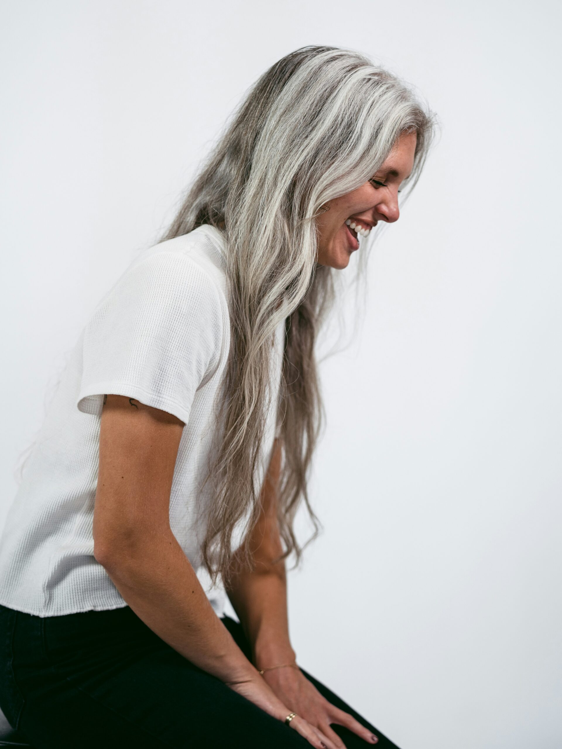 Side view of a woman sitting on a stool laughing with very long grey hair