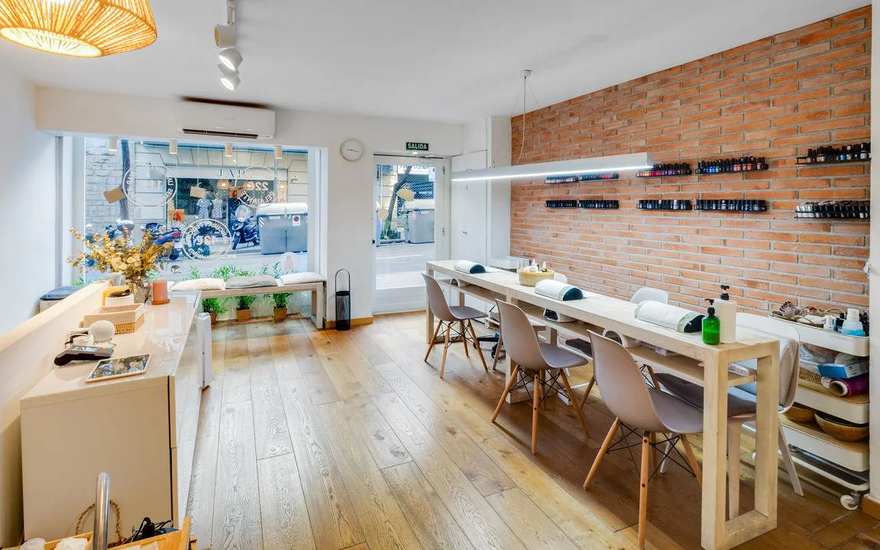 A contemporary salon with a rustic touch, featuring a long white manicure bar and a red brick accent wall. Large windows look out onto a city street, filling the wooden-floored space with natural light.