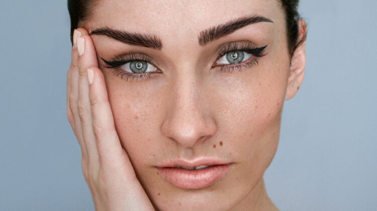 A close-up of a woman’s face with microblading on her brows and natural make-up with dark hair tied up