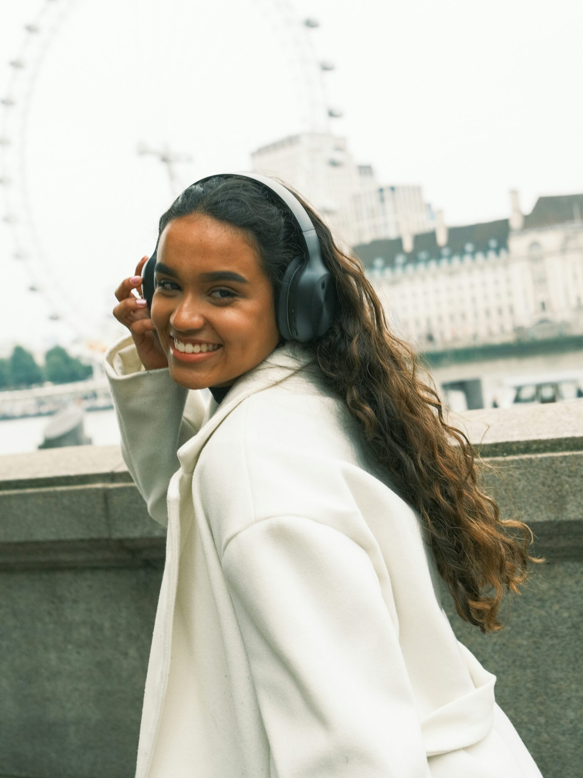 A smiling woman with microblading on her brows wearing black over-ear headphones and a white coat walking along the River Thames in London