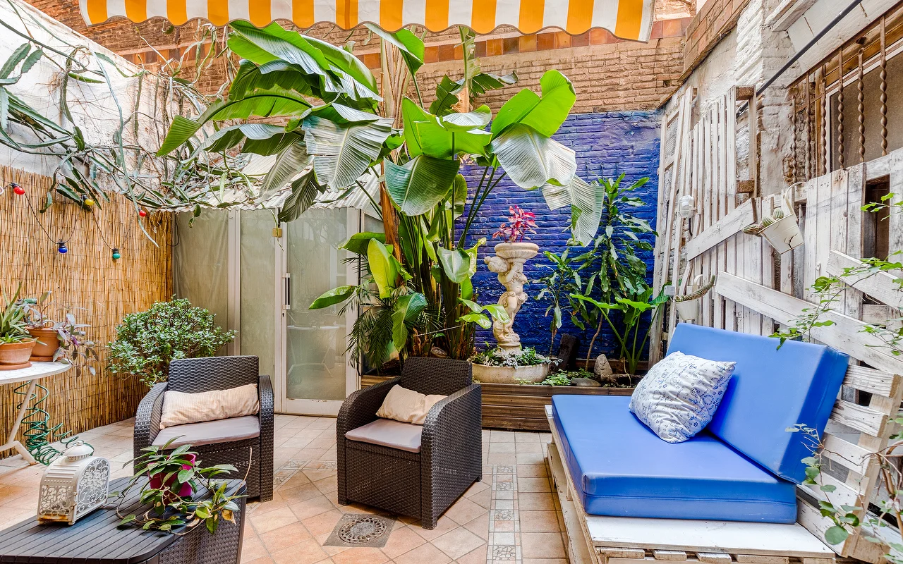 An eclectic outdoor patio or waiting area with a vibrant blue accent wall and lush tropical plants. It features dark wicker armchairs, a bright blue cushioned bench, and a yellow-and-white striped awning