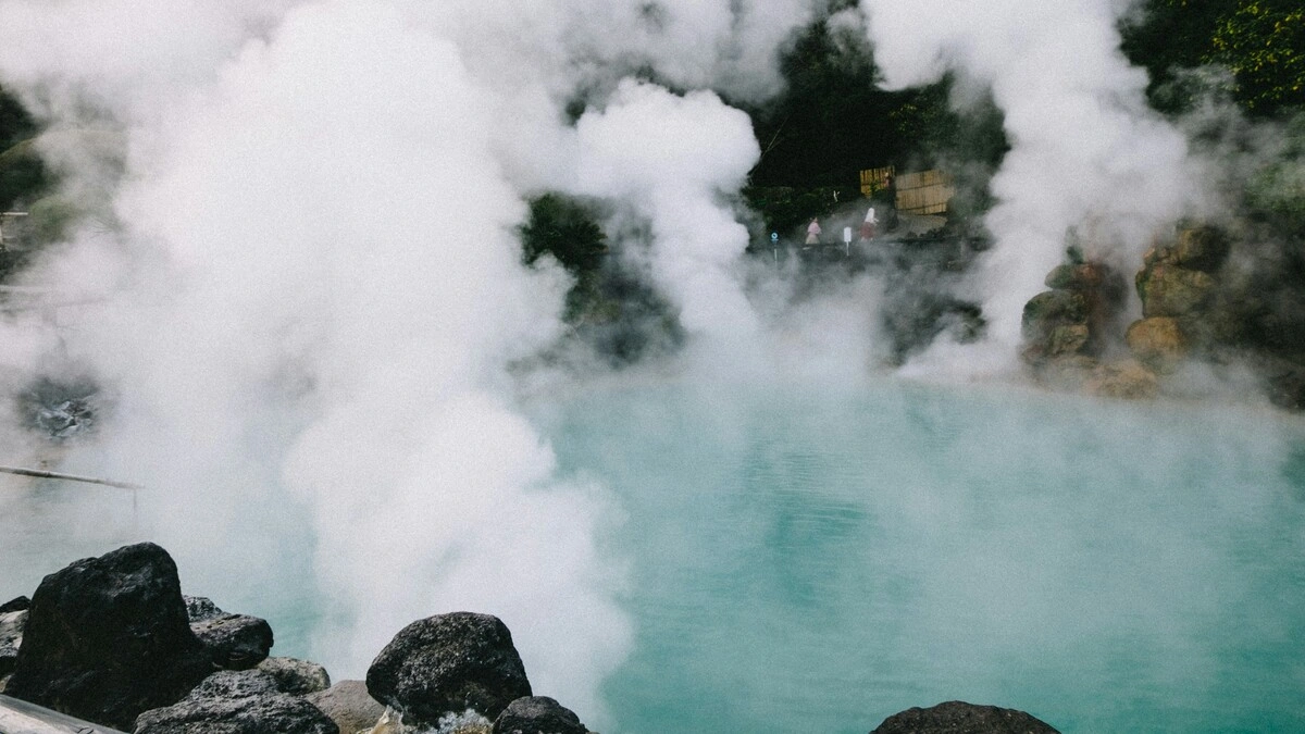 Wide view of hot springs in Japan showing clouds of steam rising from a pool surrounded by rocks and greenery