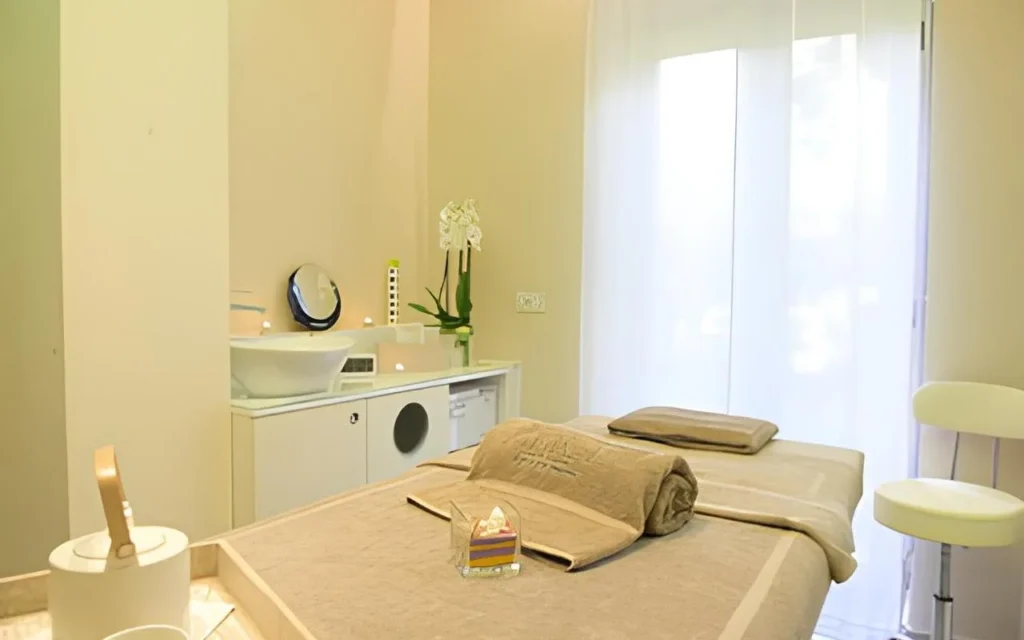 A calm treatment room at Alice Beauty Clinique with a bed dressed in soft taupe linens, white orchids on the counter, candles by the basin, and sheer curtains filtering natural light through a large window.