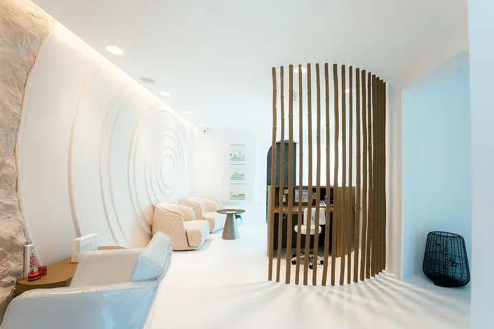 A calm, cave-like spa interior with curved white walls, one carved in concentric ripples, soft recessed lighting, cream quilted armchairs and a curved wooden-slat screen partitioning a quiet treatment nook.