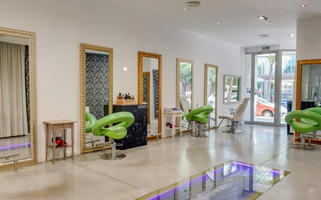 A bright, spacious hair salon with polished concrete floors, tall gold-framed mirrors along a white wall, lime-green moulded styling chairs, and a glass-floor section lit with purple light, with the street visible through glazed doors at the back.