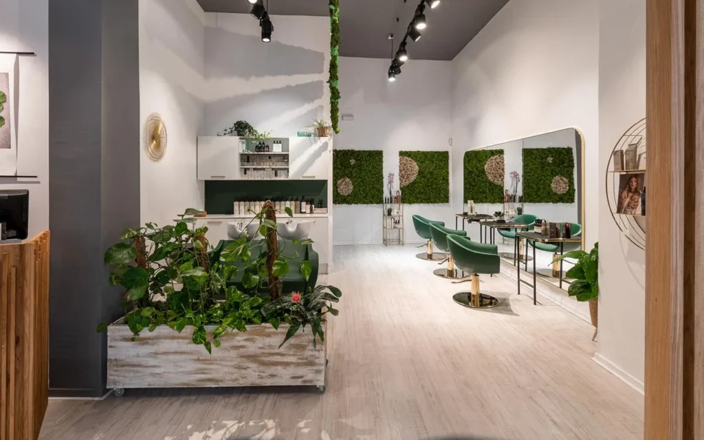A plant-filled hair salon with pale wood floors, an aged white planter overflowing with greenery, green moss wall art, emerald-green velvet styling chairs with gold detailing, and a wash station behind.