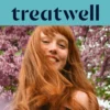 A woman with long copper-red hair and a fringe smiling in the sunshine, with pink blossom trees in the background. The Treatwell logo sits on a teal banner above.