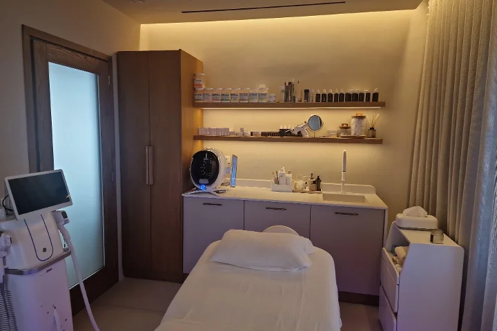 A warmly lit treatment room with a white facial bed and neatly folded towels, dark wood shelving lined with skincare products, a skin-analysis screen and treatment machines alongside, clinical but cosy.