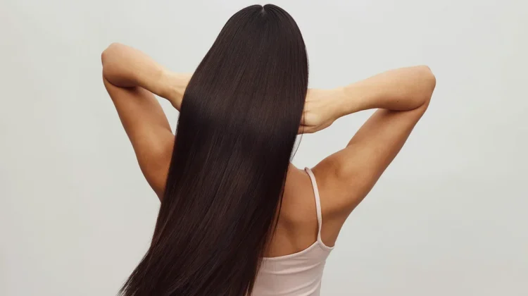 Back view of a woman wearing a white strappy top with her arms up running her fingers through her long, glossy brunette hair