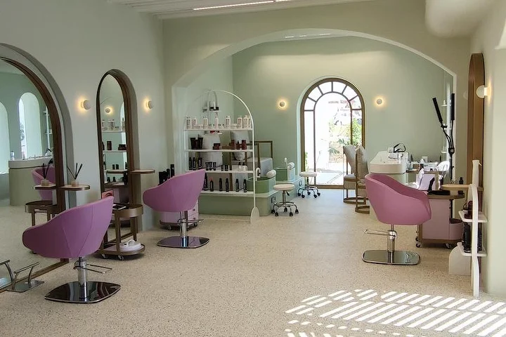 A bright sage-green salon with terrazzo flooring, arched wood-framed mirrors and an arched window to the garden, dusky-pink styling chairs and open shelves of products, with sunlight falling across the floor.