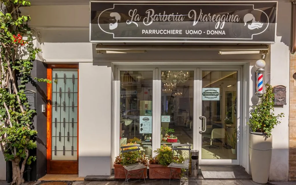 The storefront of La Barberia Viareggina on a sunny day, with a classic red, white and blue barber pole to the right of the glass doors. Potted plants and climbing greenery frame the entrance, and a chandelier is visible inside through the windows.