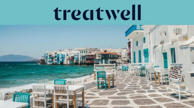 The Little Venice waterfront in Mykonos Town: whitewashed houses with colourful wooden balconies at the sea's edge, the turquoise Aegean beyond, and white and teal café tables set out across a stone-paved terrace in the foreground.