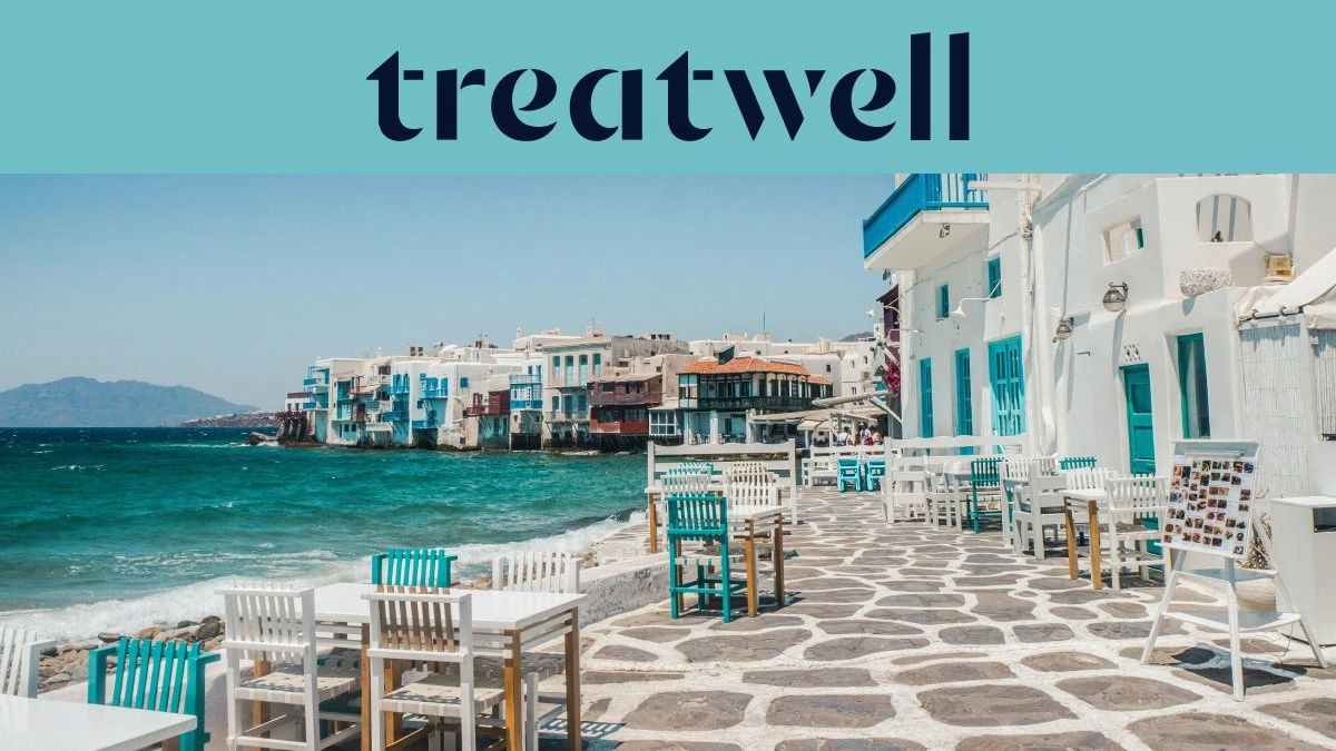 The Little Venice waterfront in Mykonos Town: whitewashed houses with colourful wooden balconies at the sea's edge, the turquoise Aegean beyond, and white and teal café tables set out across a stone-paved terrace in the foreground.