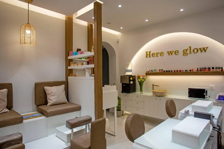 A warm, softly lit nail salon with a woven rattan pendant lamp, cushioned brown pedicure benches, white manicure tables, a shelf of colourful polishes and a gold 'Here we glow' sign on a backlit arch.