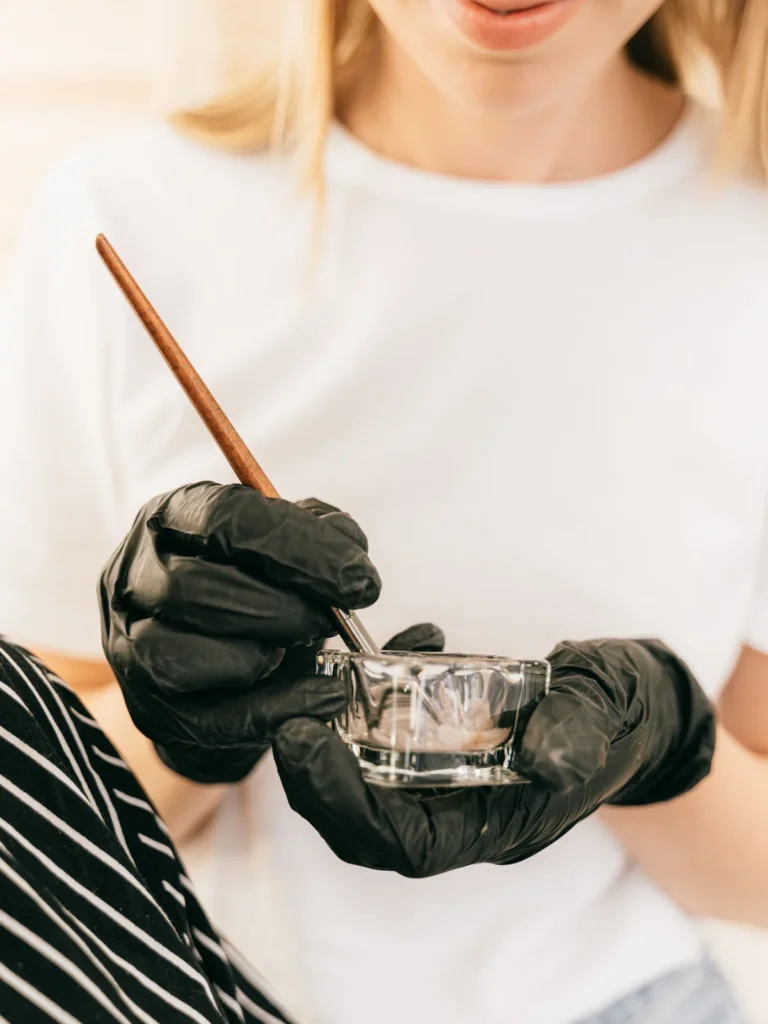 Close-up of a technician holding a small glass pot and a thin brush, mixing eyebrow tint in a salon