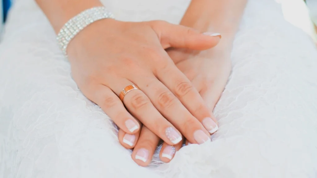 A woman’s hands rest on her lap, she is wearing a lace wedding dress and has a neat French tips manicure with a classic nude and white combination on short squoval nails and delicate embellishment on her ring fingers