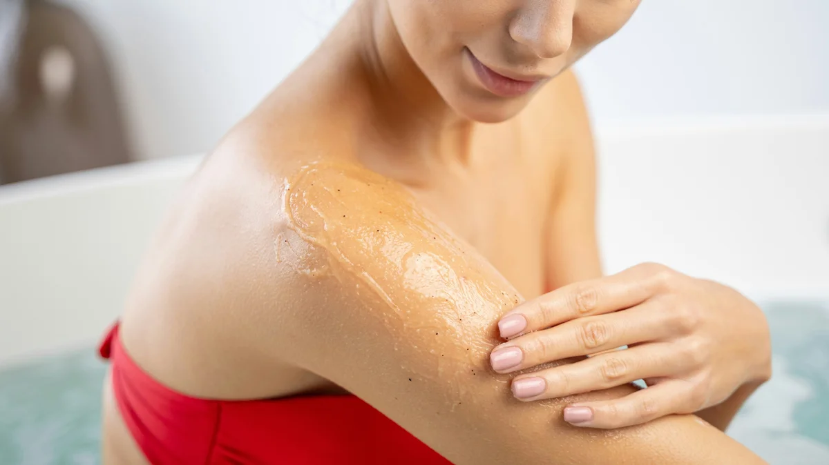 Close-up of a woman wearing a red bikini and applying exfoliating gel to her upper arm in a clean, white bathroom