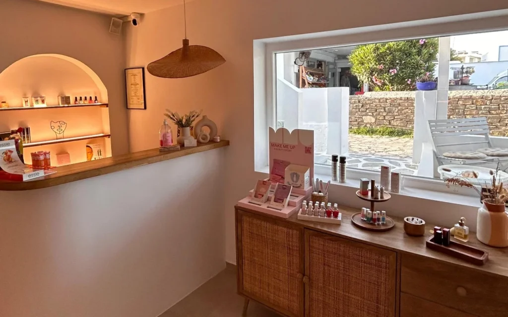 The interior of BeautiQ Esthetics and More on Paros, with a softly lit arched niche of nail varnishes, a woven rattan pendant and a large window onto a stone wall and sunlit garden.