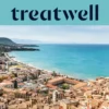 A sweeping view over a Sicilian coastal town, with terracotta-roofed old buildings, a crescent of sandy beach, clear turquoise sea and a rocky headland rising to one side.