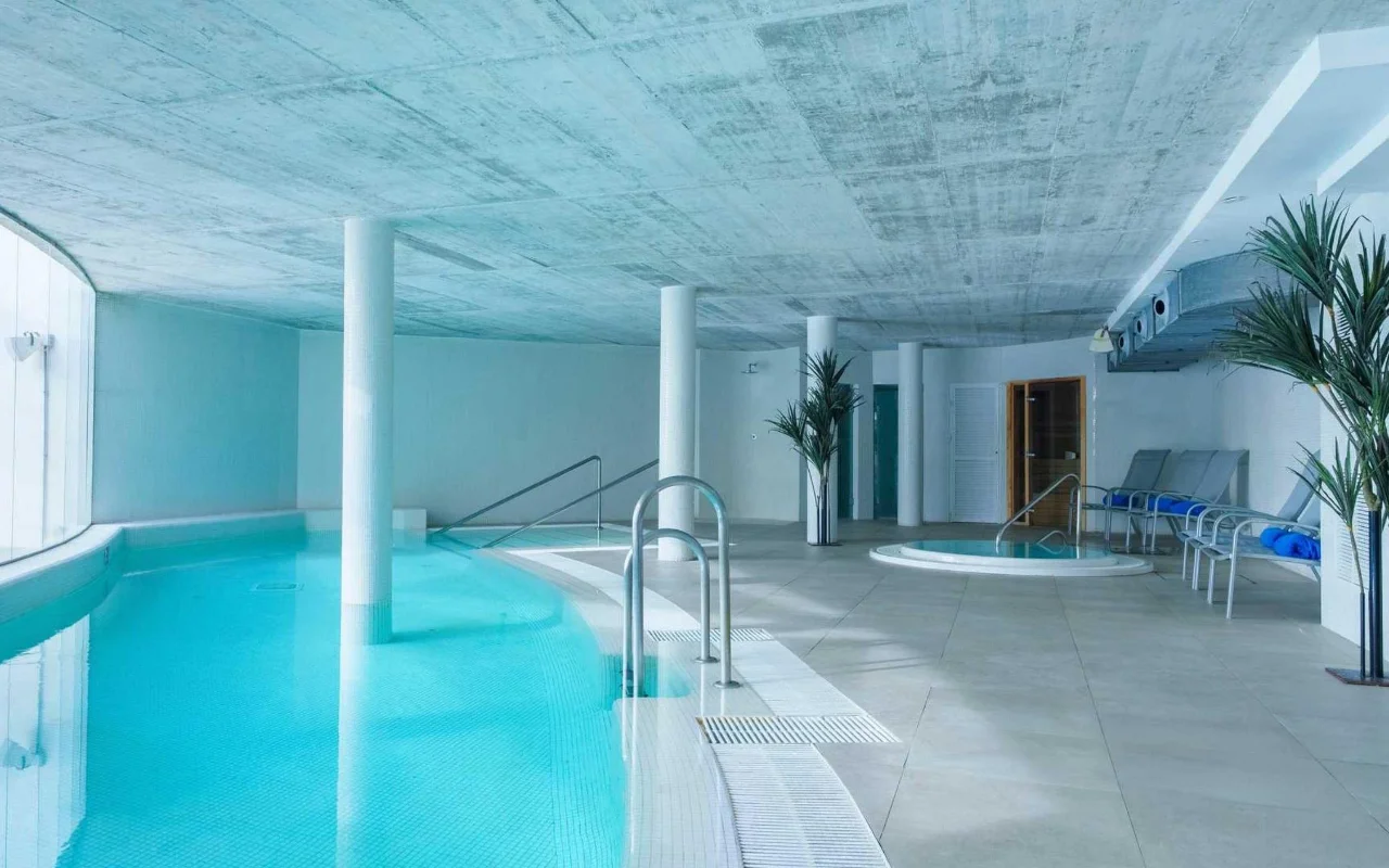 The indoor spa pool at Benalmádena Beach, with a curved turquoise pool, an exposed concrete ceiling, a small jacuzzi and loungers among tall potted palms.