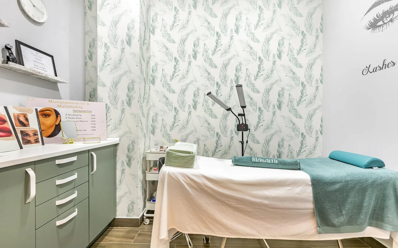 A treatment room at Masami Belleza Natural in Fuengirola, with a therapy bed, feather-patterned wallpaper, a magnifying lamp and a micropigmentation price display.