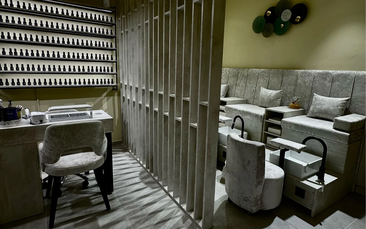 A treatment room at Sisu Nail Club and Academy in Perissa, Santorini, with a full wall of nail varnishes, grey velvet manicure and pedicure chairs and a concrete slatted screen.