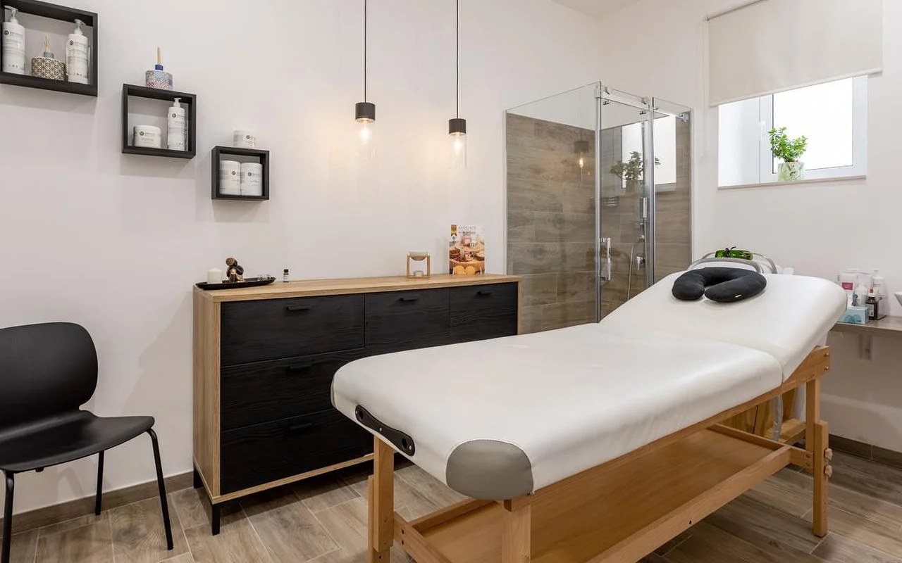 A massage room at Thea Estetica, with a wooden massage table topped by a black face cradle, a black-and-wood chest of drawers, two pendant lights, box shelves of products and a glass shower cubicle with wood-effect tiling.