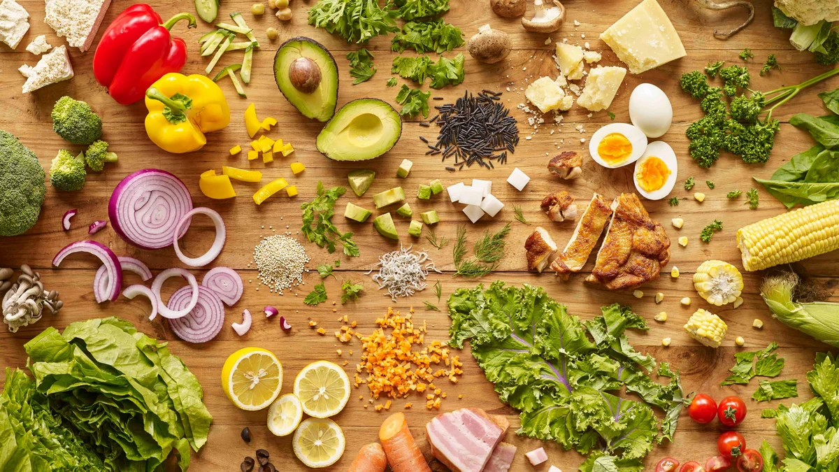 Top view of a wooden kitchen counter covered in bright and colourful nutritious food, including eggs, vegetables, salad and chicken