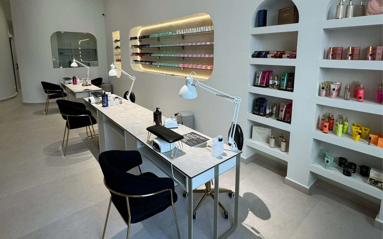 A white nail studio with marble-topped manicure tables and white anglepoise lamps, black velvet chairs on gold legs, and arched wall niches displaying skincare and a backlit rack of nail polishes.