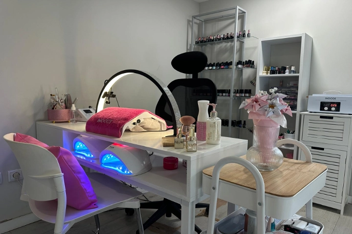 A single nail station with a white desk, a ring light and two blue-glowing LED lamps, a black chair and shelves stacked with colourful nail polishes, brightened by a pink vase of flowers.