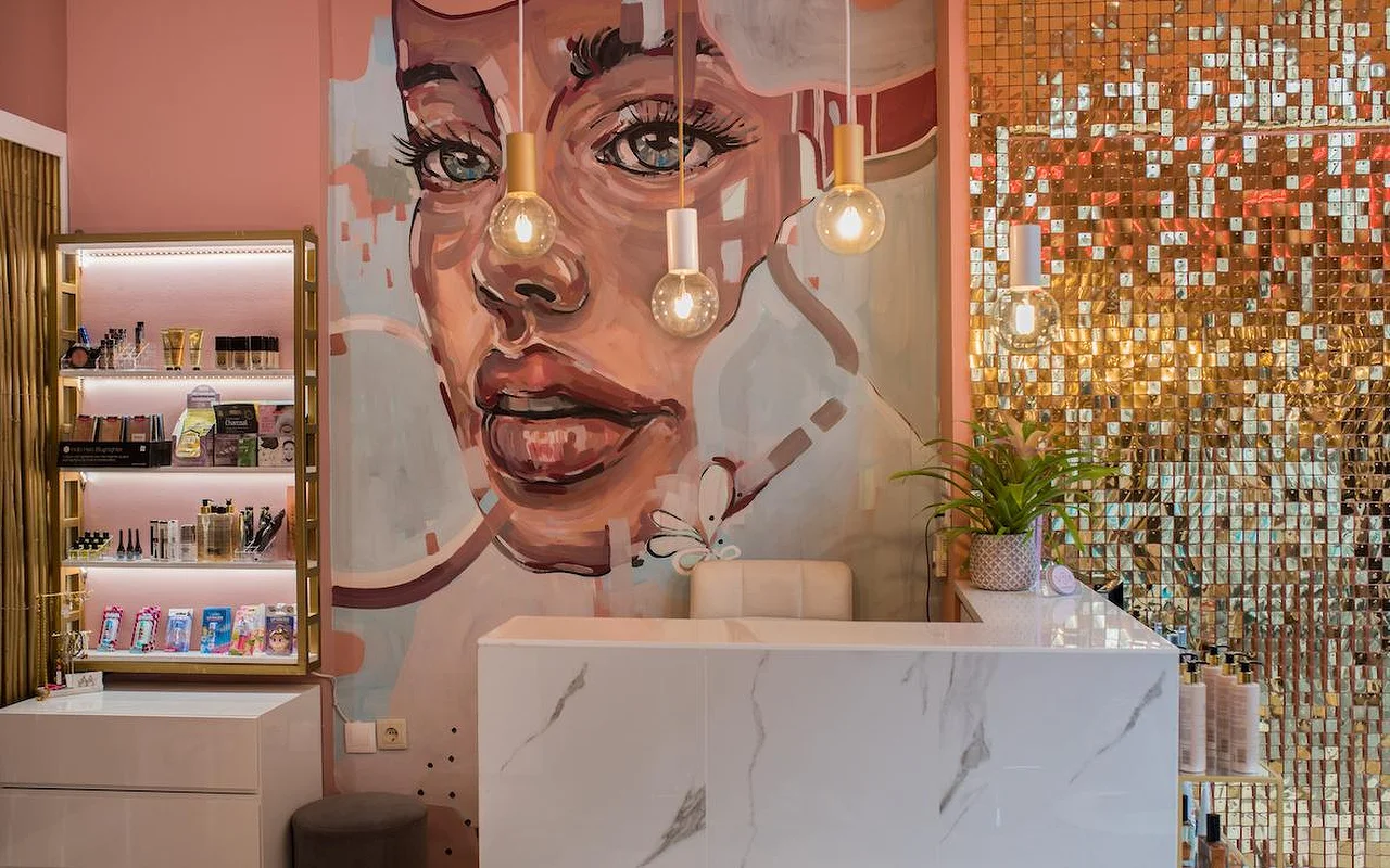 A pink-walled salon with a large painted mural of a woman's face, a shimmering gold sequin wall, a white marble reception counter and bare-bulb pendant lights.